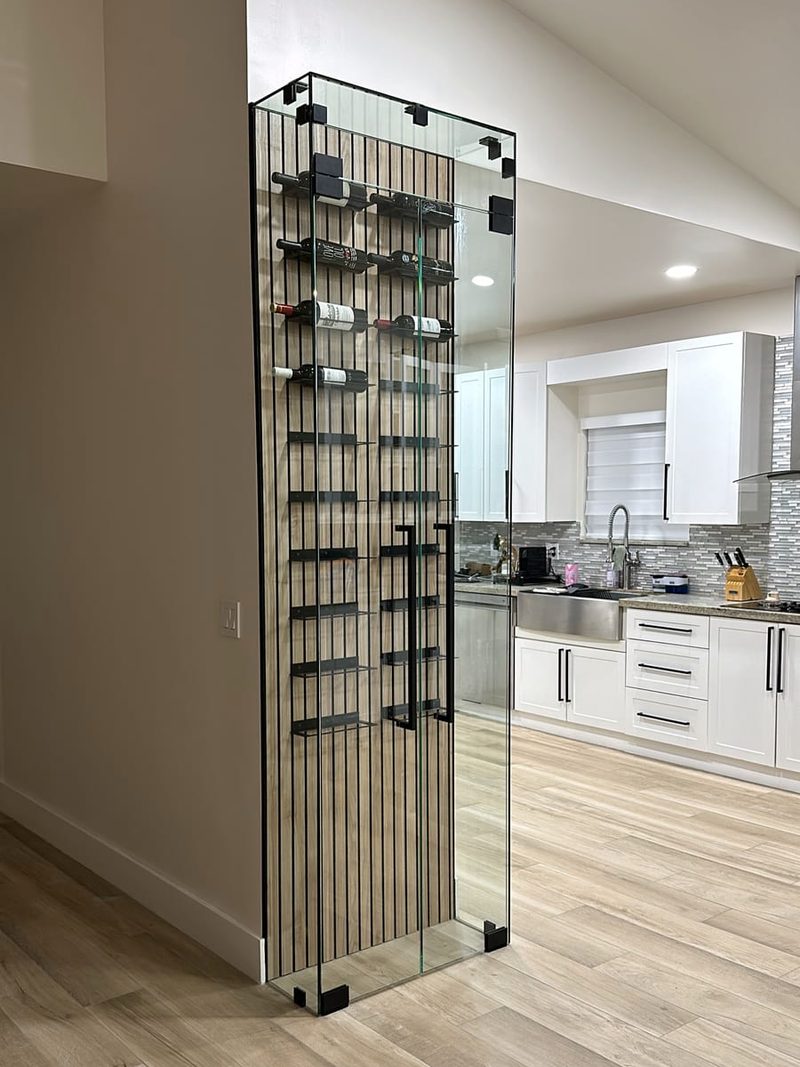 Floor-to-ceiling glass wine display enclosure with black corner clamps and a black pull handle, next to a kitchen