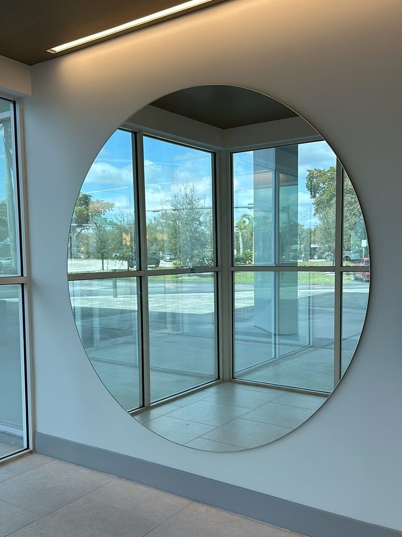 Large round frameless mirror on a lobby wall reflecting floor-to-ceiling windows