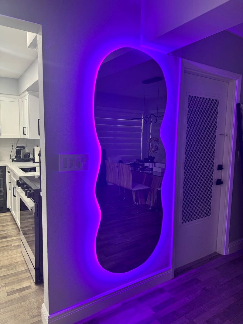 Tall wavy-edge mirror with purple LED backlighting in a hallway next to a kitchen