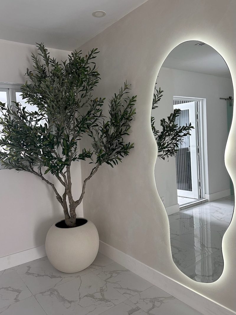 Tall wavy-edge mirror with LED backlight on a white wall next to an olive tree in a round planter