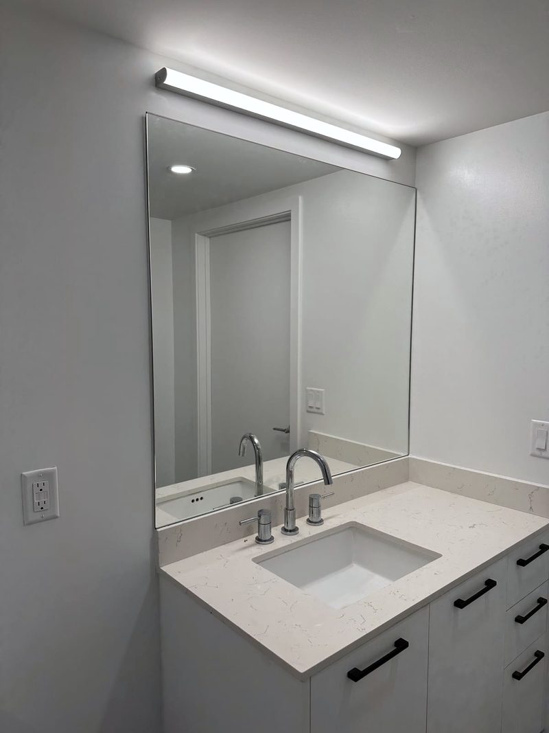 Large frameless rectangular bathroom mirror above a white quartz vanity with a light bar above