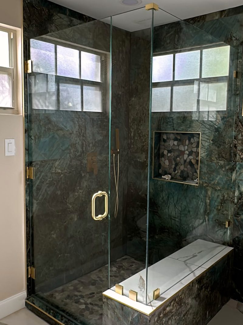 Frameless glass shower enclosure with brushed gold hinges, clamps and handle over dark green stone walls and a bench