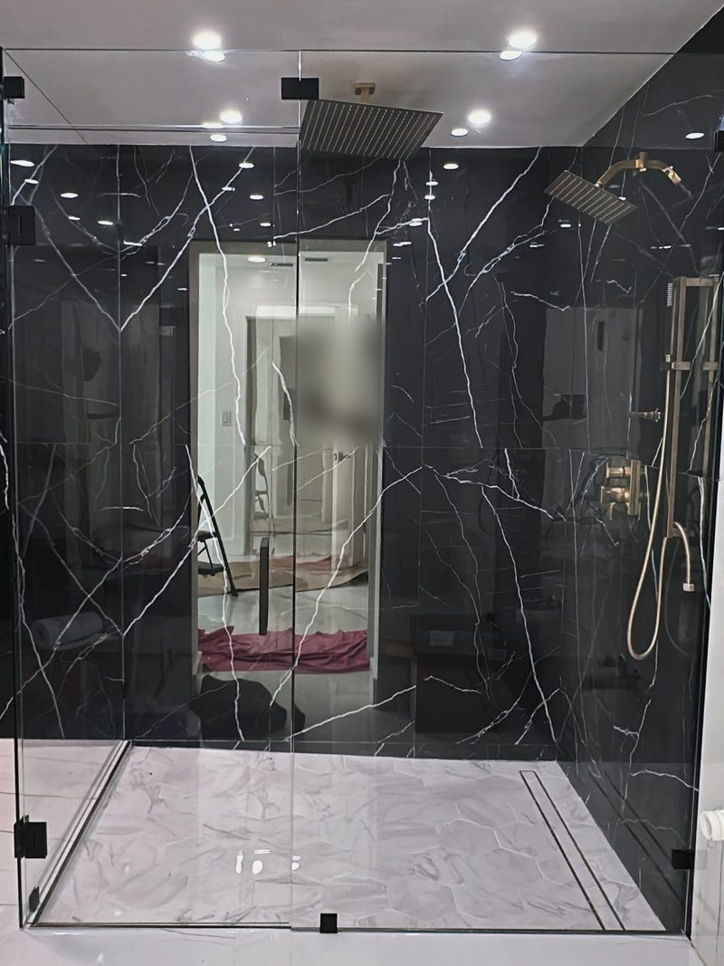 Frameless glass shower enclosure with black clips against black marble walls, with brushed gold fixtures